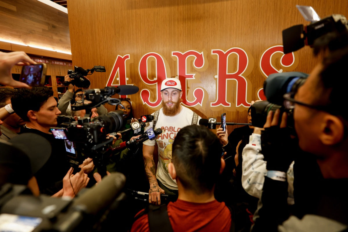George Kittle in an All American STALLY hat surrounded by reporters and cameras with '49ers' branding in the background