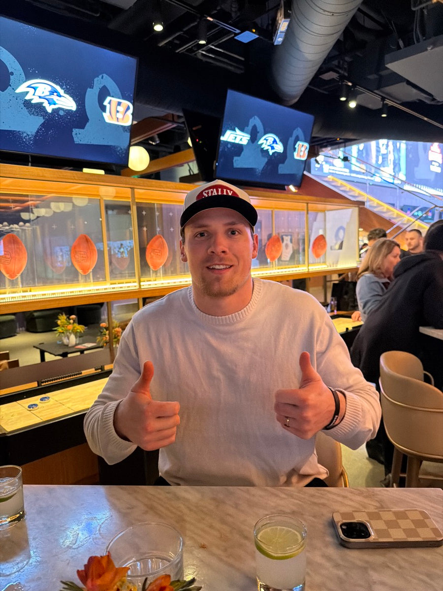 Jack Campbell in a white sweater and All American STALLY hat standing in a restaurant with a glass display case in the background.