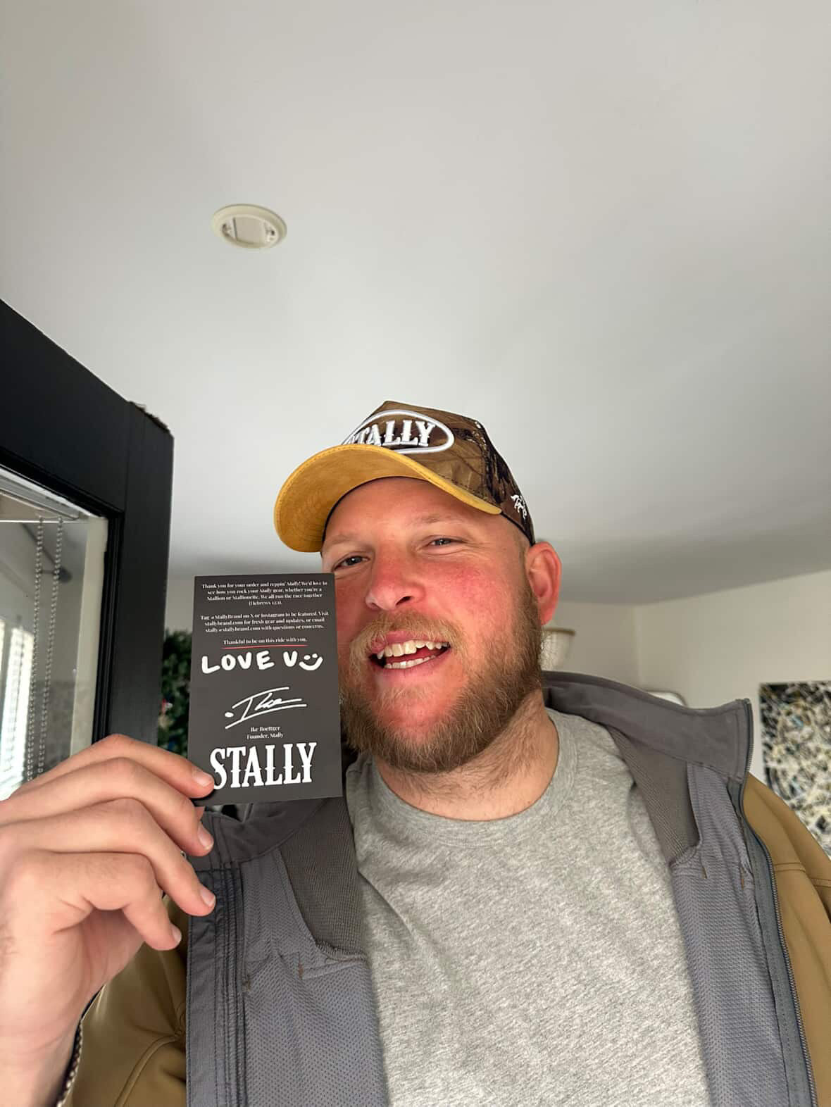 Man wearing a CAMO STALLY hat and holding a product with 'STALLY' branding indoors.