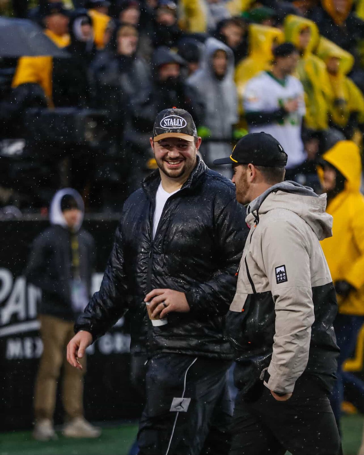 Two men - Ike Boettger and Josey Jewell - walking on Kinnick Stadium in the rain, wearing winter clothing and CAMO STALLY hats.