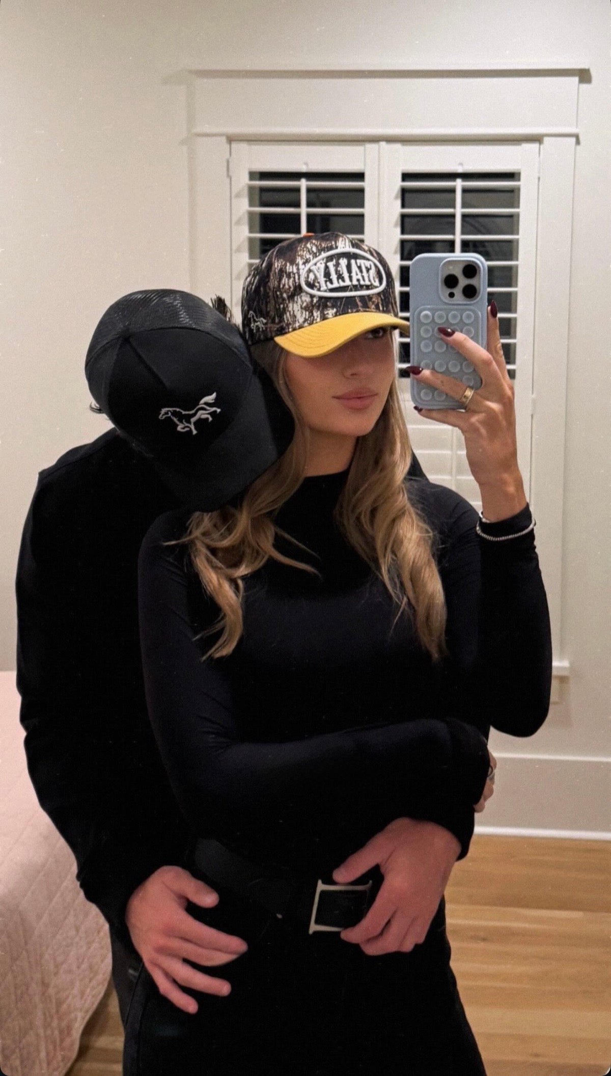 Two people wearing black hoodies and STALLY hats taking a mirror selfie.