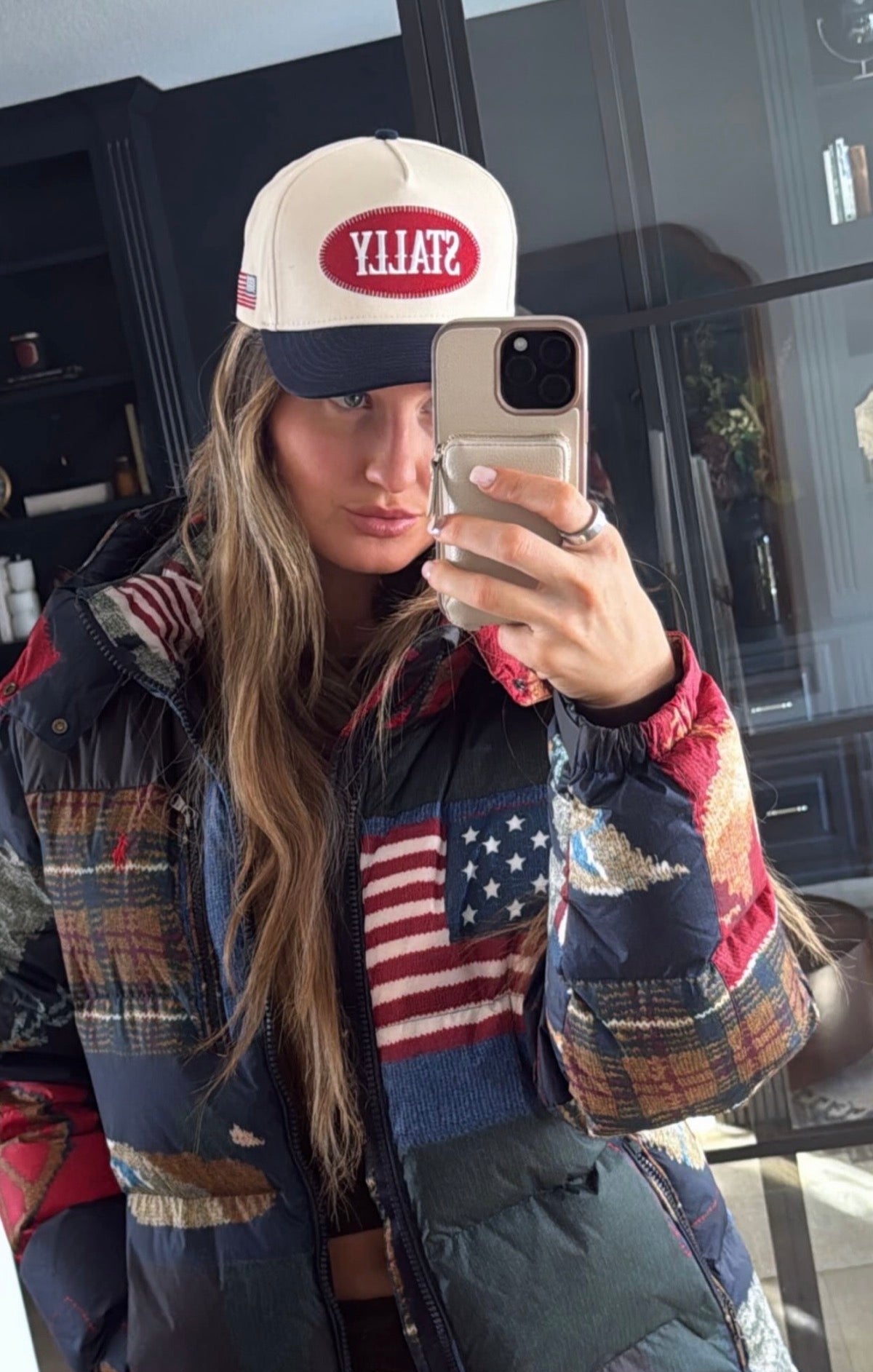 Person wearing an All American STALLY hat and a patchwork jacket with American flag design, taking a mirror selfie.