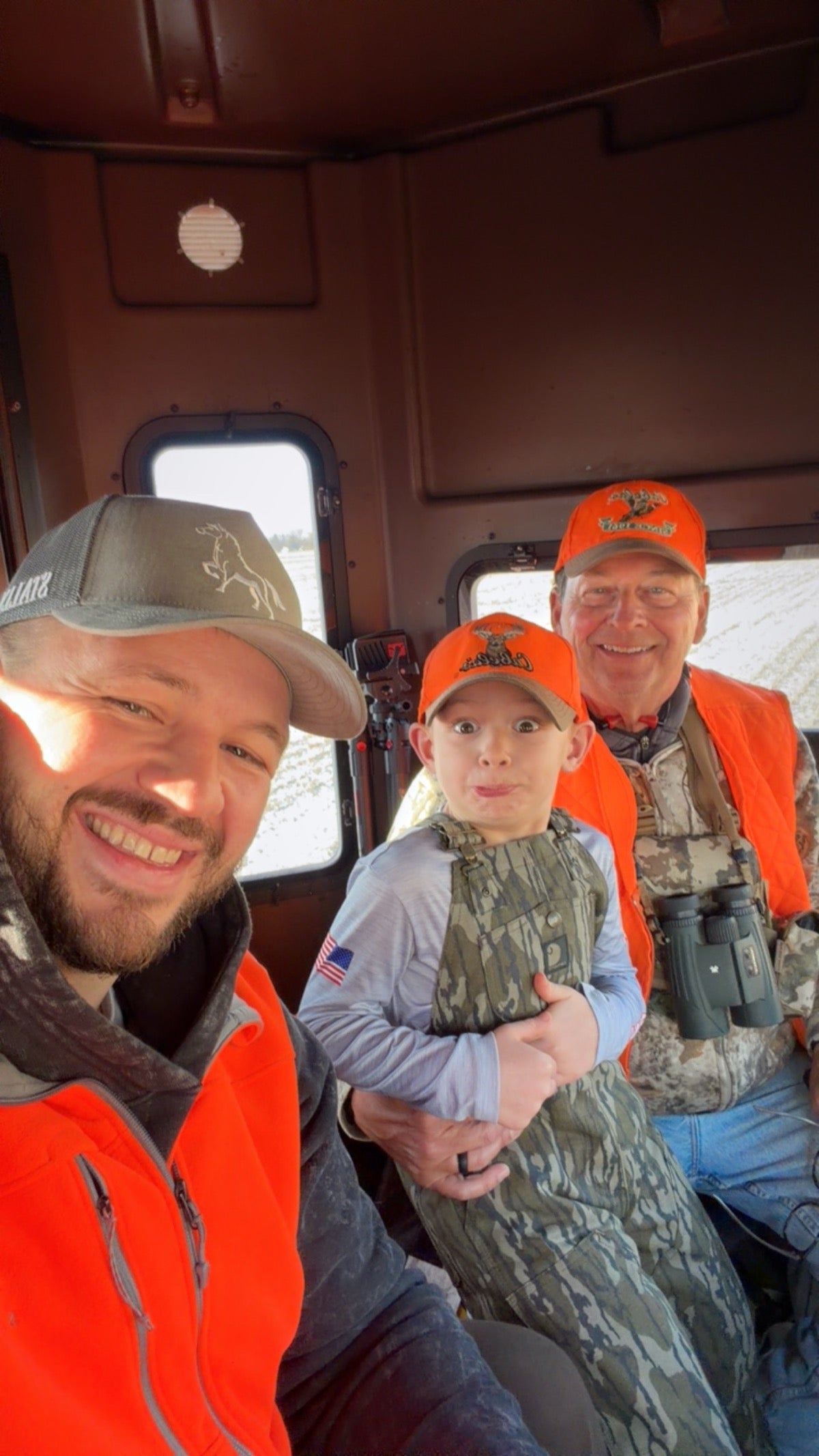 Three people, two adults and a child - one adult wearing a Charcoal OG STALLY hat, sitting inside a vehicle wearing hunting gear.