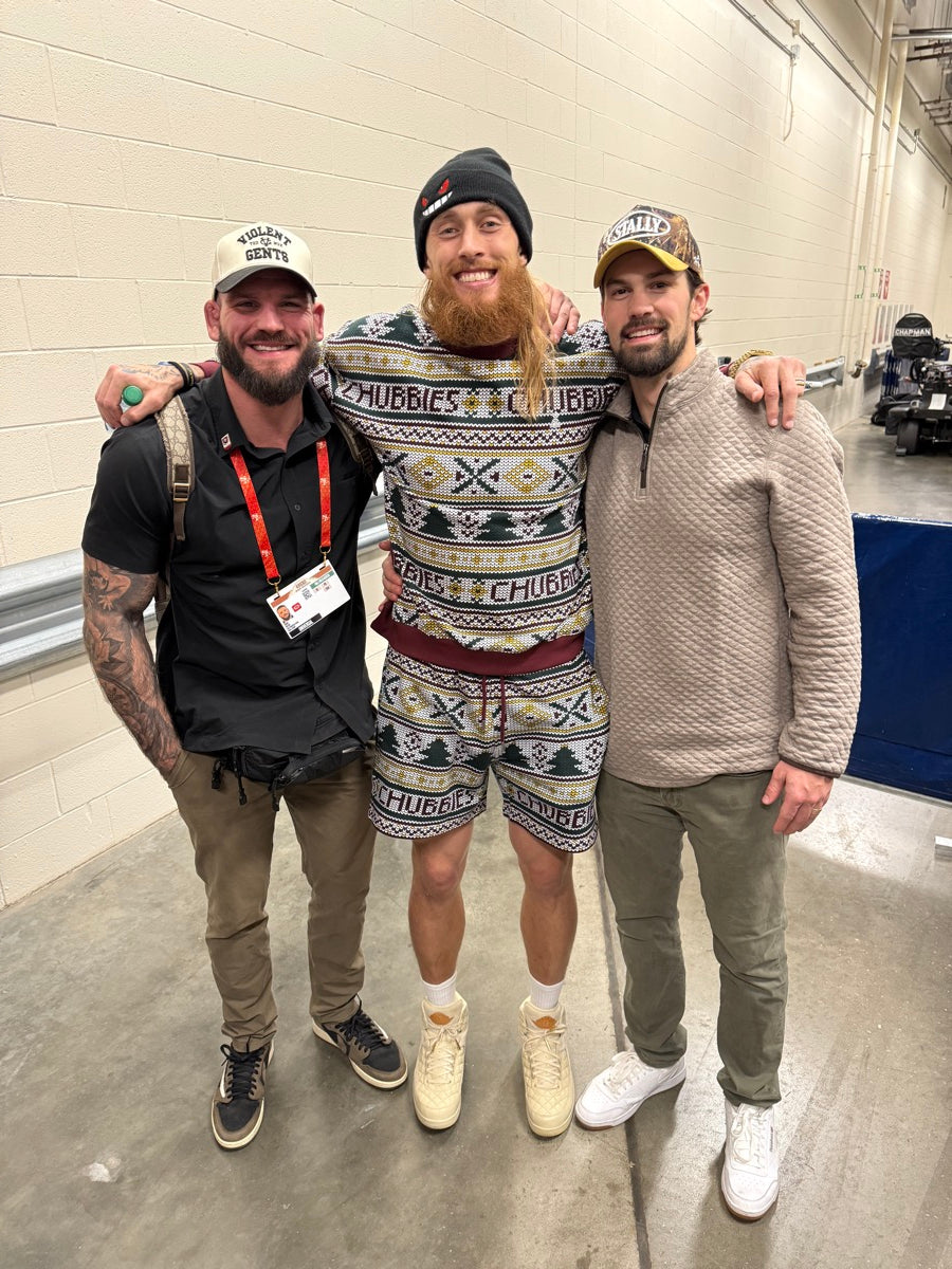 Three people posing together in a hallway with a neutral background, one wearing a CAMO STALLY hat