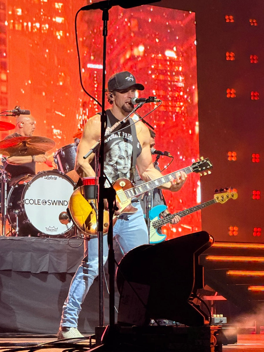 Musicians performing with Cole Swindell on stage with instruments and a red lighted background with the singer wearing an OG STALLY hat