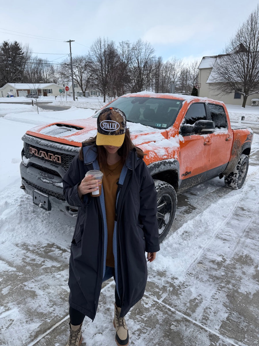 Person wearing a CAMO STALLY hat standing next to a snow-covered vehicle on a snowy street