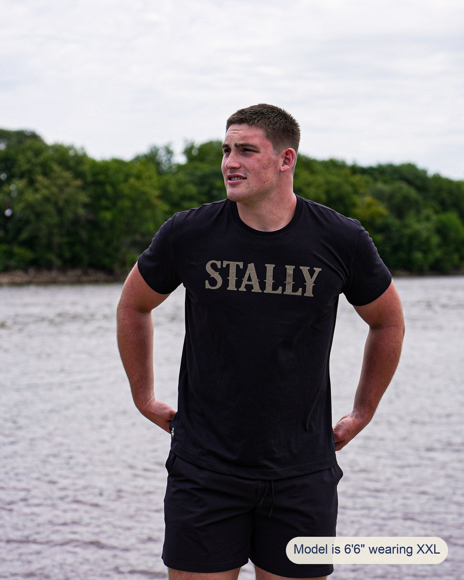 A man standing outdoors by the water wearing a black t-shirt with 'STALLY' printed across the chest in a distressed gold font, paired with black shorts. A text overlay at the bottom right reads 'Model is 6'6" wearing XXL'.