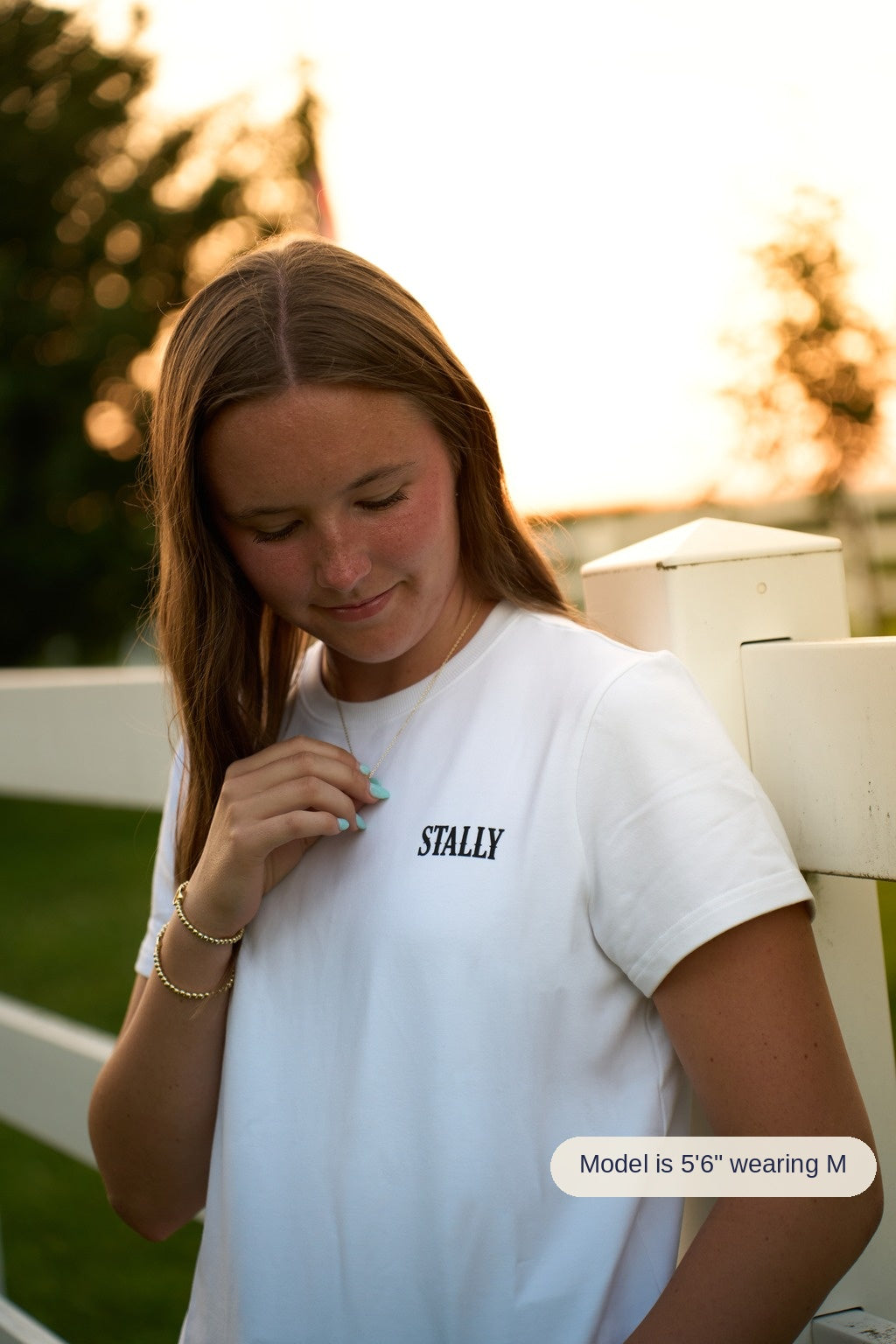 A woman leans against a white fence at golden hour, looking down while wearing a white crewneck t-shirt with 'STALLY' printed on the chest, a gold necklace, and bracelets.