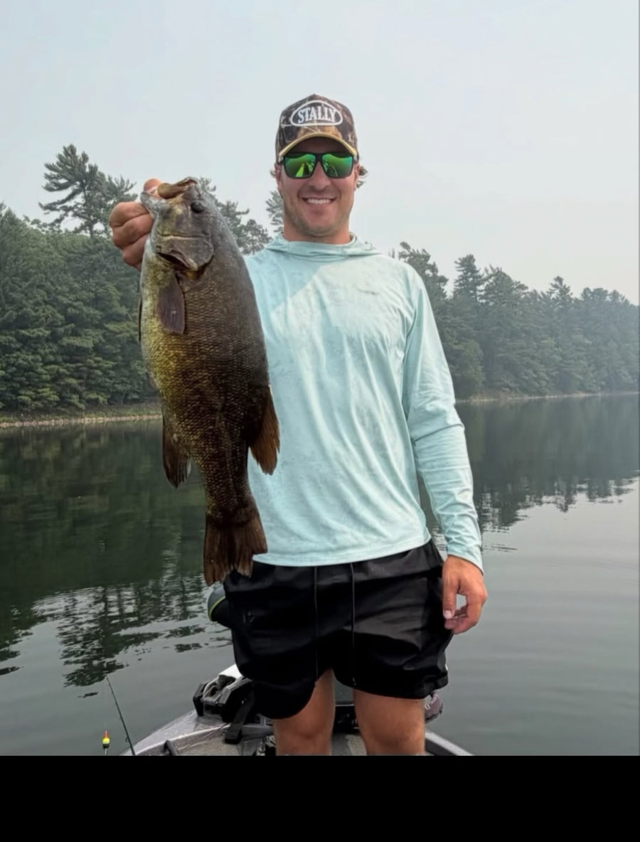 An angler in a Stally camo cap holding a fish beside a lake.
