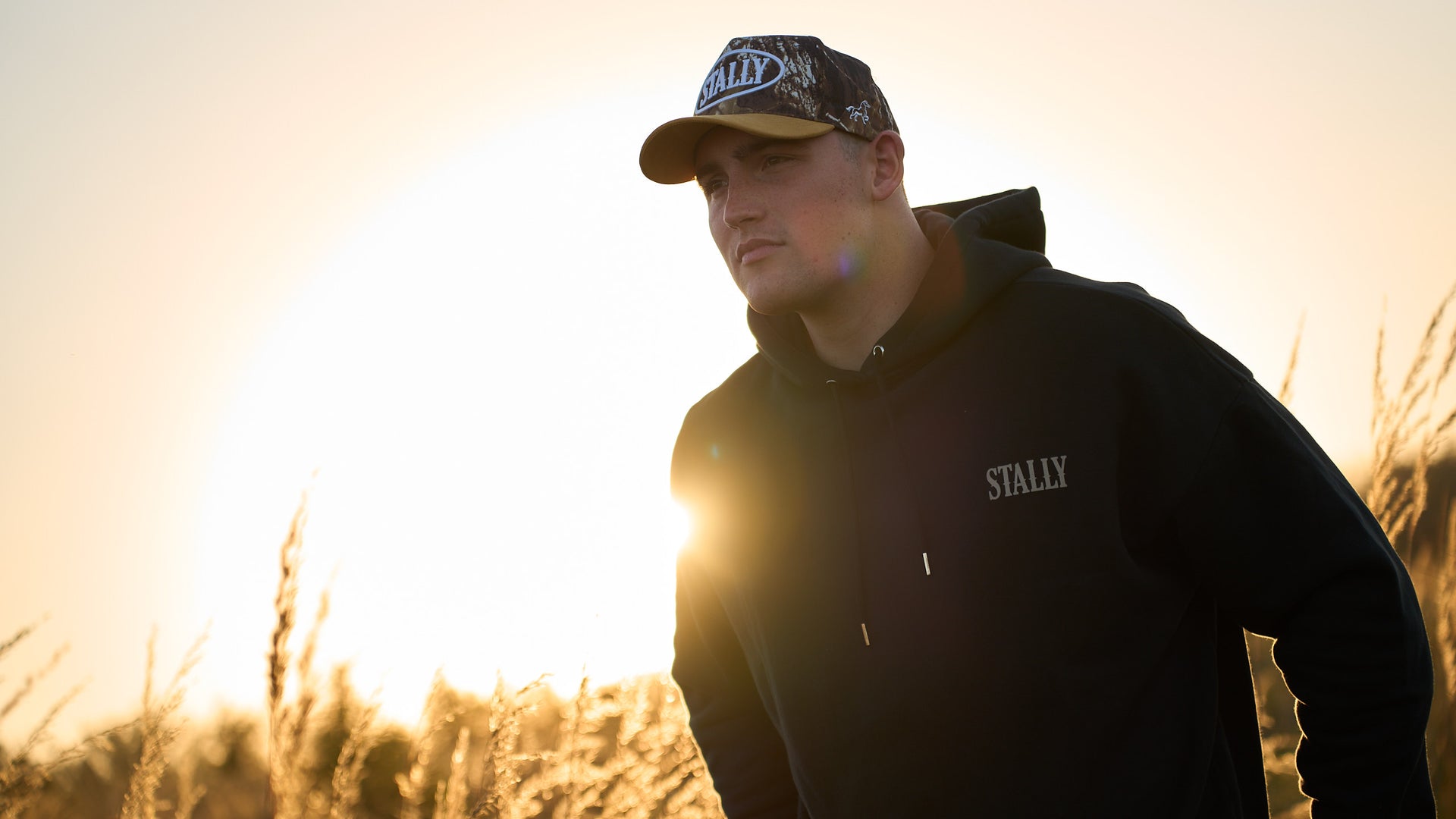 A man in a Coal Black Stally Draft hoodie and a camo oval-patch cap in tall grass at sunset