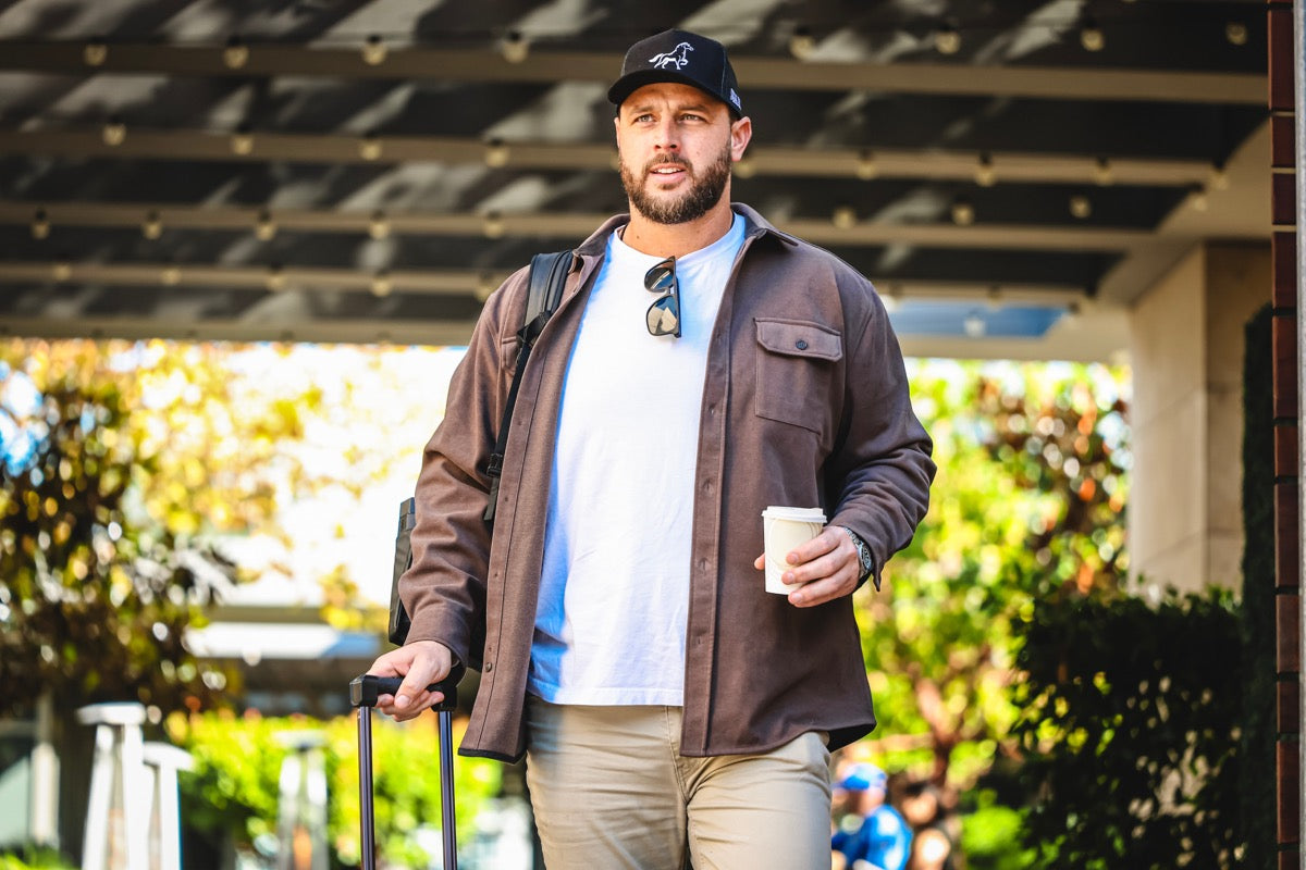 NFL player wearing an OG STALLY hat with a suitcase and coffee cup walking outdoors on his way to a game