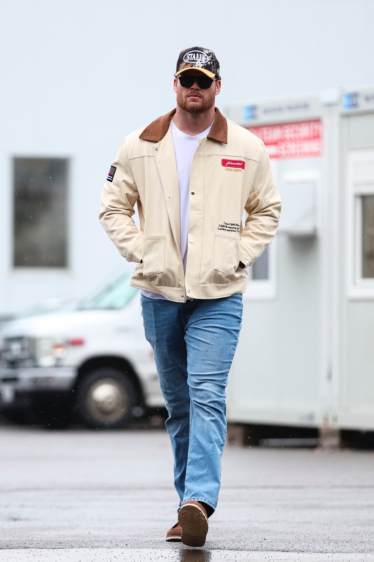 Man - Spencer Brown - wearing a beige jacket with patches, blue jeans, and a CAMO STALLY hat walking on a street to play a game for the Buffalo Bills.