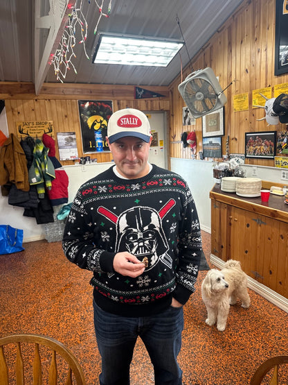 Person wearing an All American STALLY hat and a festive sweater with a Darth Vader design in a room with wooden floors and various items.