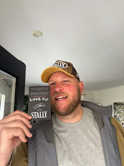 Man wearing a CAMO STALLY hat and holding a product with 'STALLY' branding indoors.