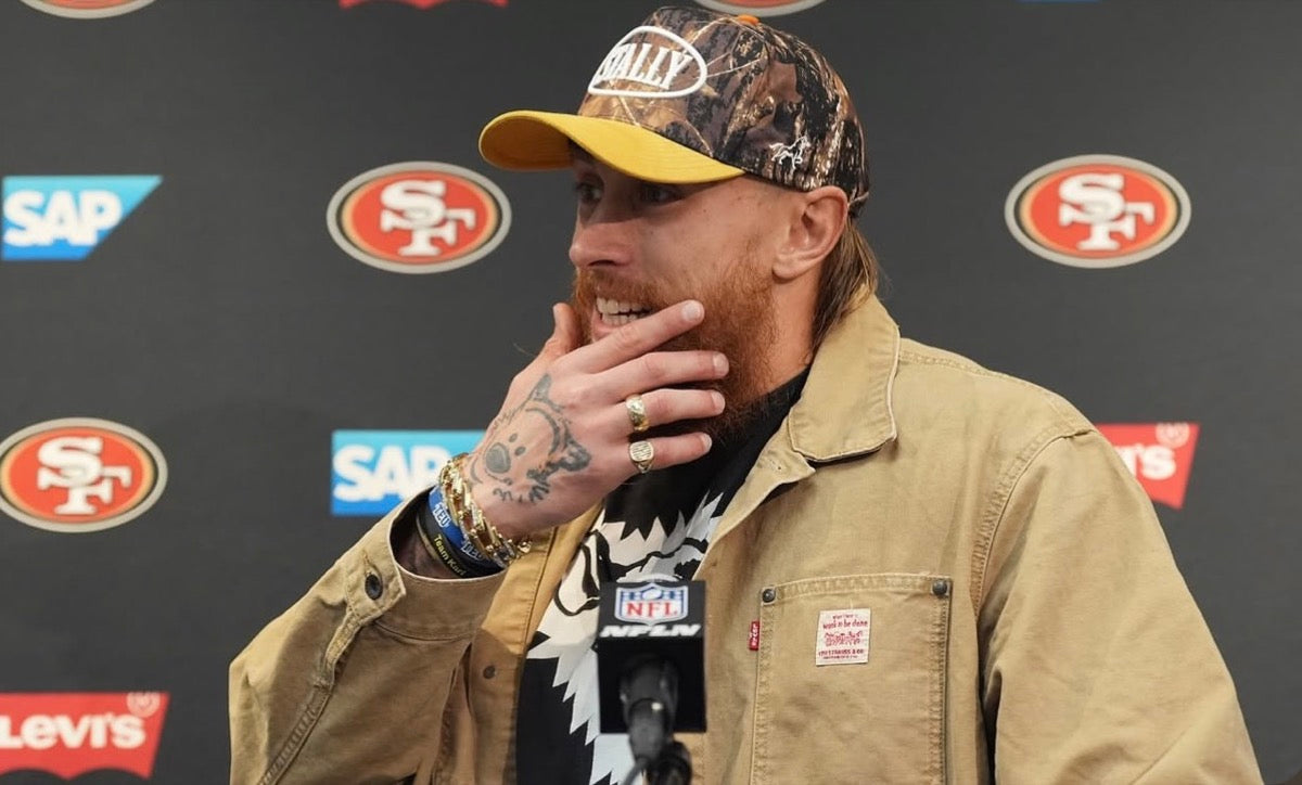 George Kittle wearing a CAMO STALLY hat at a press conference with 49ers logos in the background
