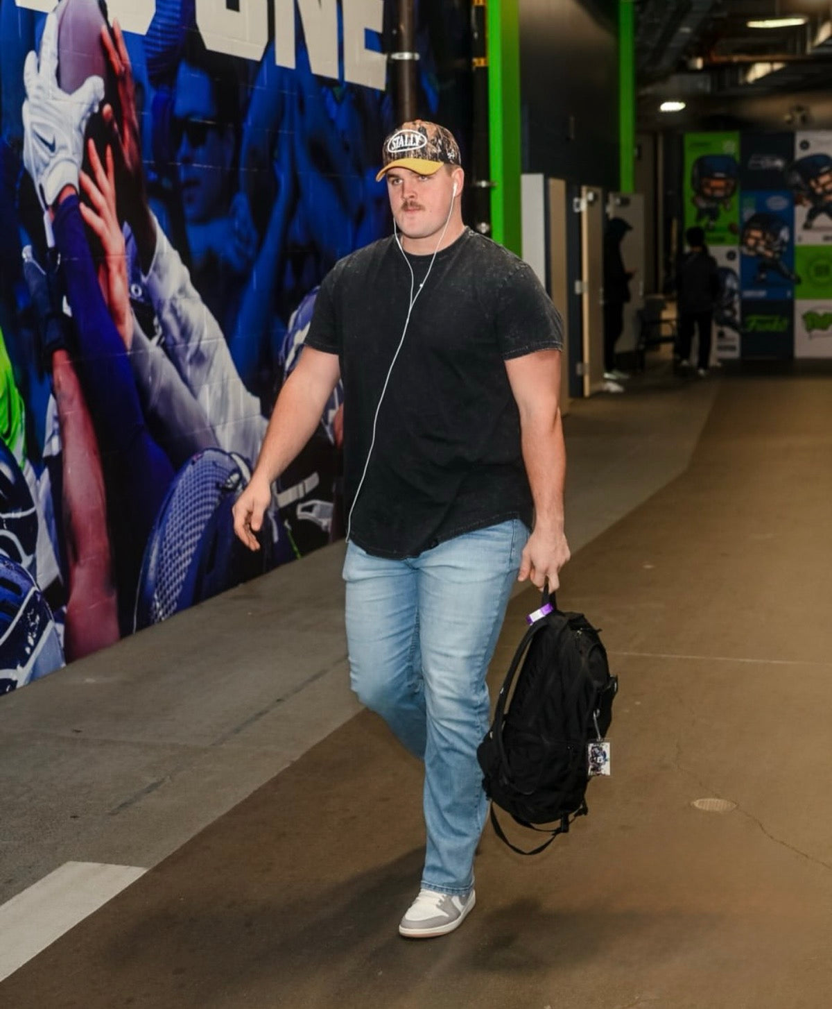 Grey Zabel wearing a CAMO STALLY hat walking with a backpack in an indoor setting with a colorful mural in the background. On his way to play for the Seahawks