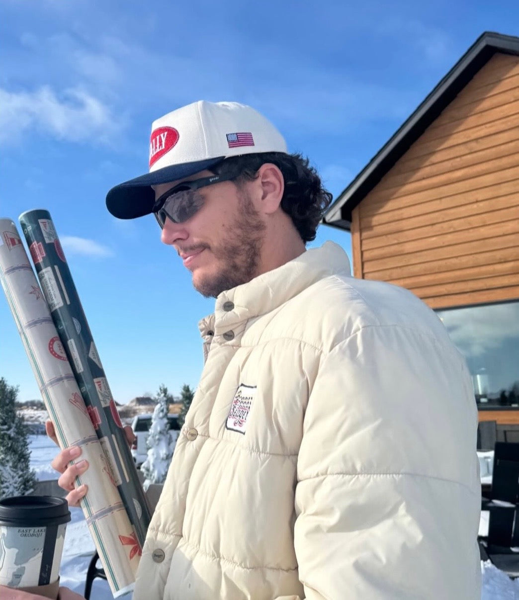 Man in winter clothing wearing an All American STALLY hat holding ice fishing rods with a cabin in the background