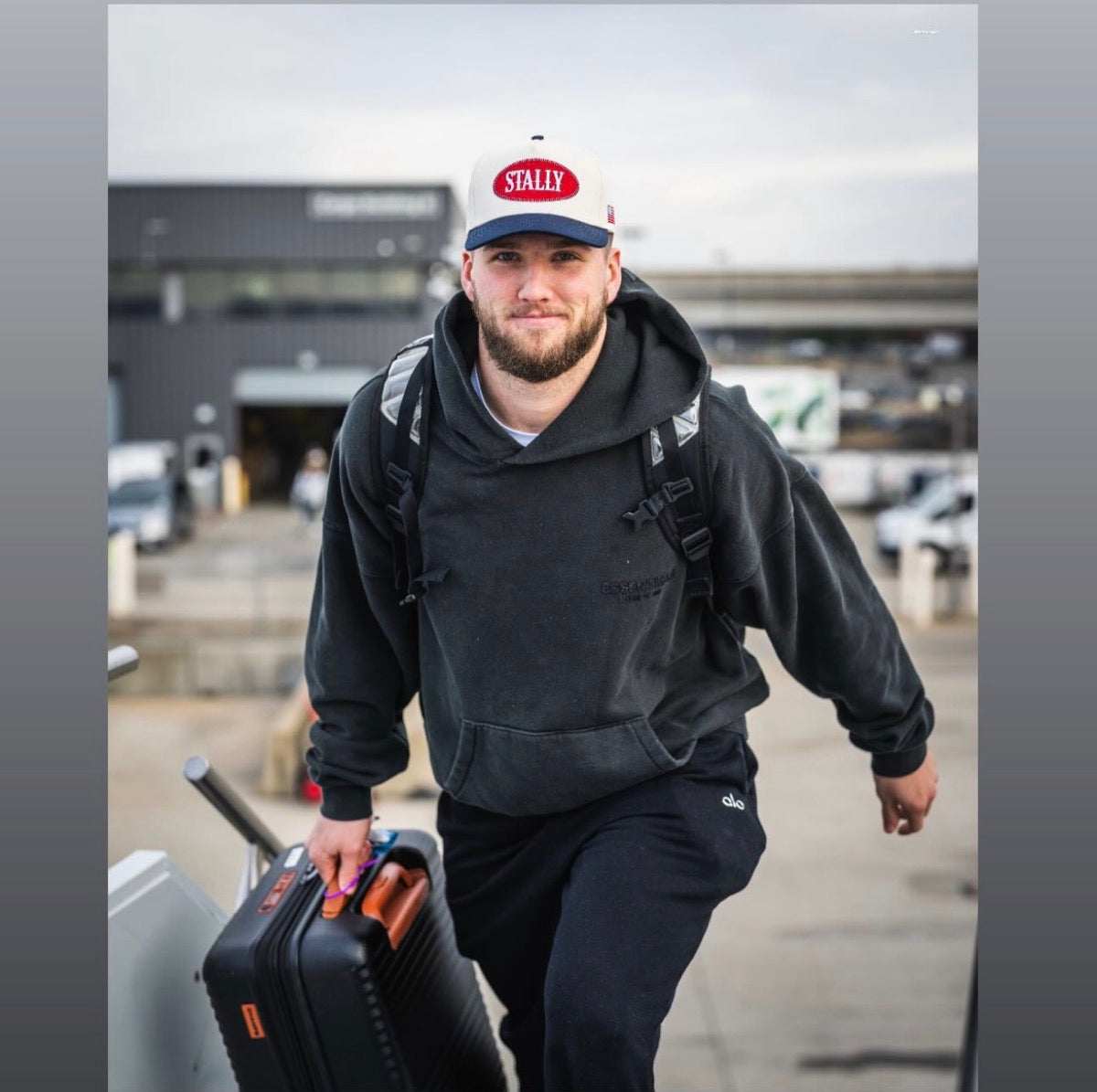 Ben Sinnott in a black hoodie and All American STALLY hat, carrying a suitcase at an airport.