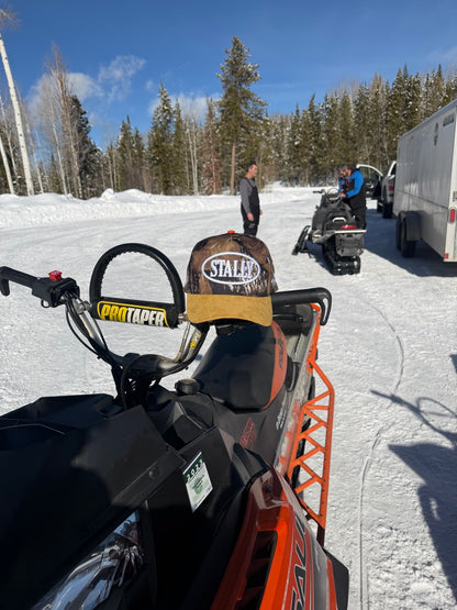 Snowmobile with a CAMO STALLY hat on a snowy landscape with trees and a truck in the background.