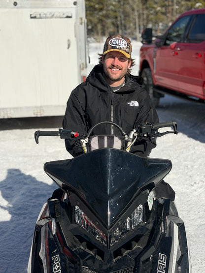 Man sitting on a snowmobile wearing a CAMO STALLY hat in a snowy landscape with vehicles in the background