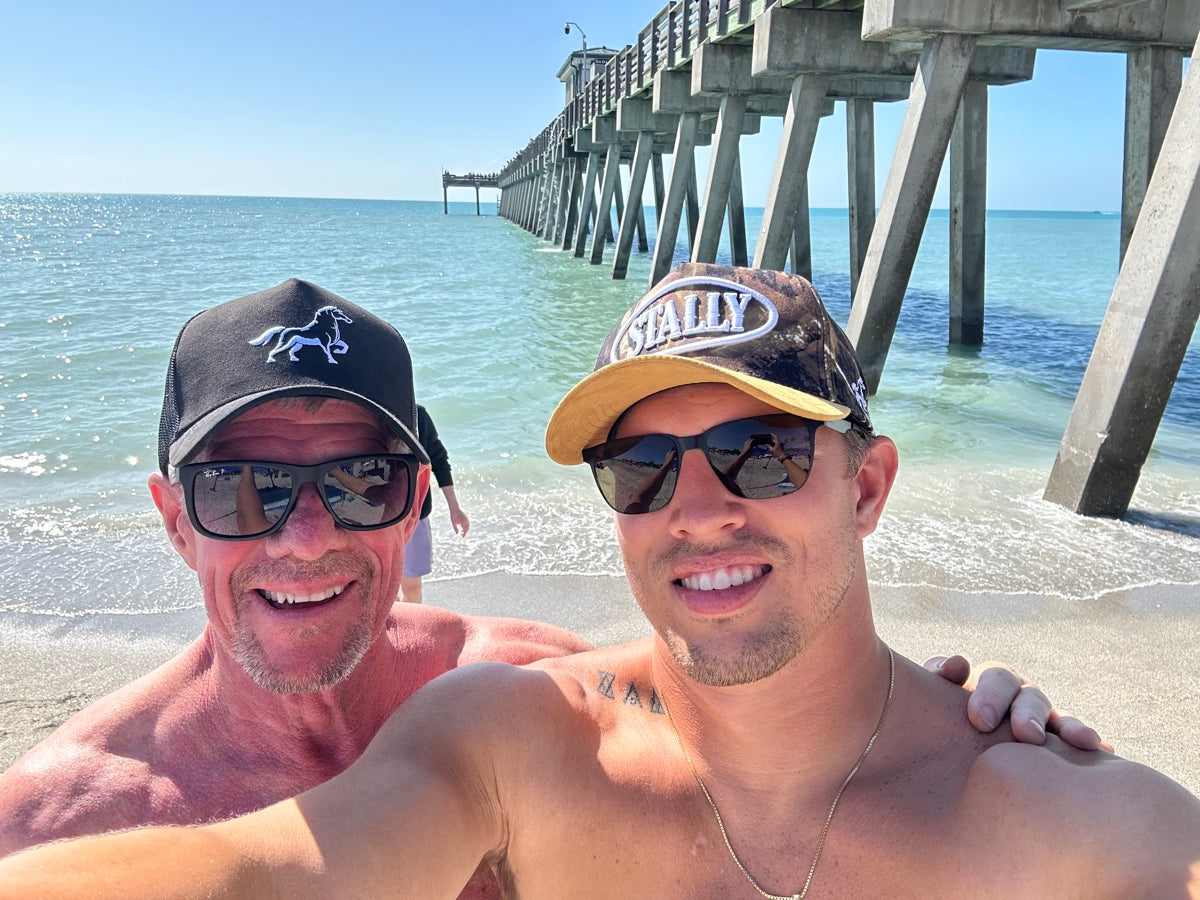 Two people wearing sunglasses and STALLY hats with a pier and ocean in the background