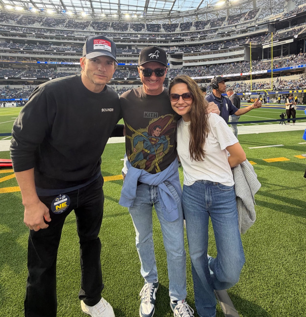 Three people - one is Ashton Kutcher - one wearing an OG STALLY hat - posing on a nfl football field with stadium in the background