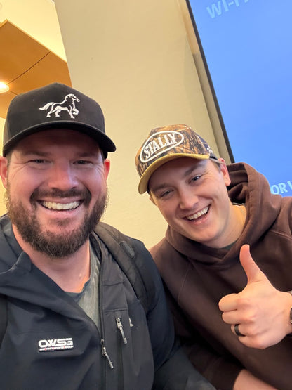 Two men wearing STALLY hats posing for a selfie outdoors with a clear blue sky