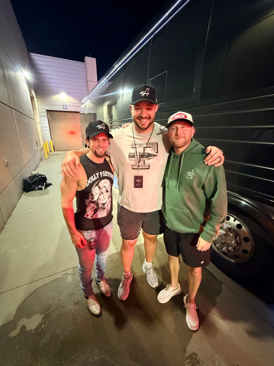 Three people wearing STALLY hats standing together in a garage with a car in the background