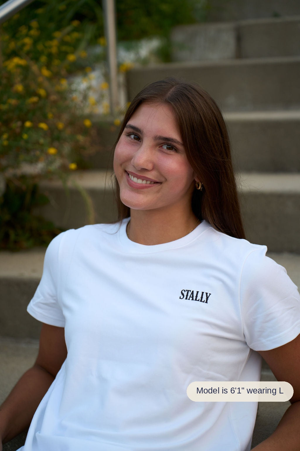 A smiling young woman with long brown hair is seated outdoors on concrete steps, wearing a white short-sleeve T-shirt with the brand name 'STALLY' printed in black on the left chest.