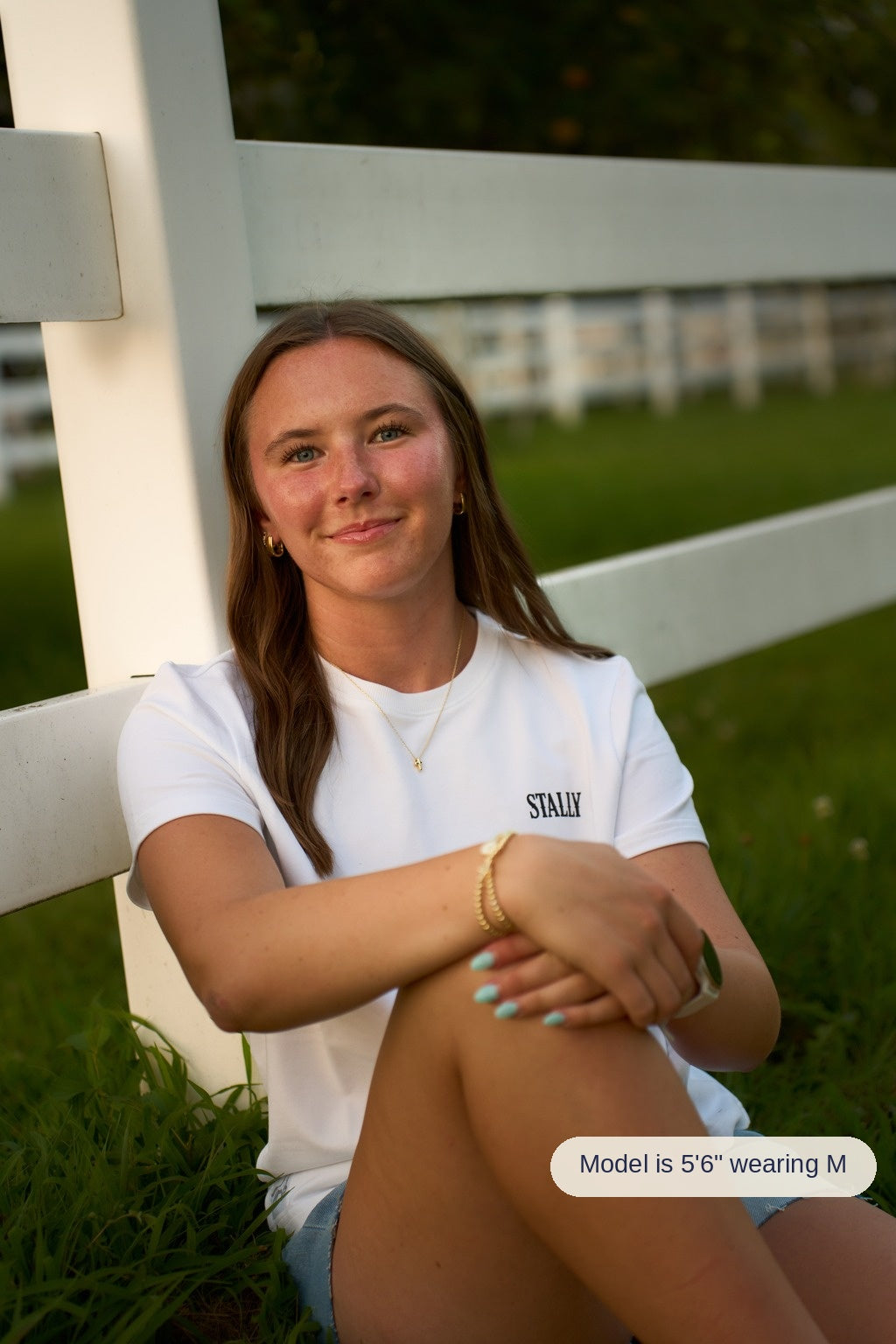 A smiling woman with long brown hair sits against a white fence rail in front of a grassy field, wearing a white crewneck t-shirt with 'STALLY' printed on the chest.