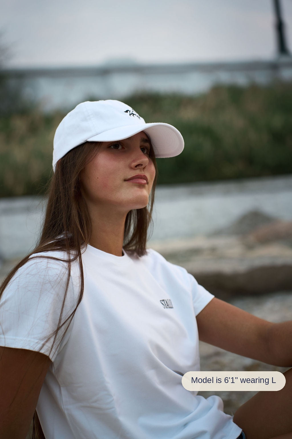 A woman wearing a white baseball cap and a white t-shirt with a small black logo, photographed outdoors near a body of water with a blurred natural background.