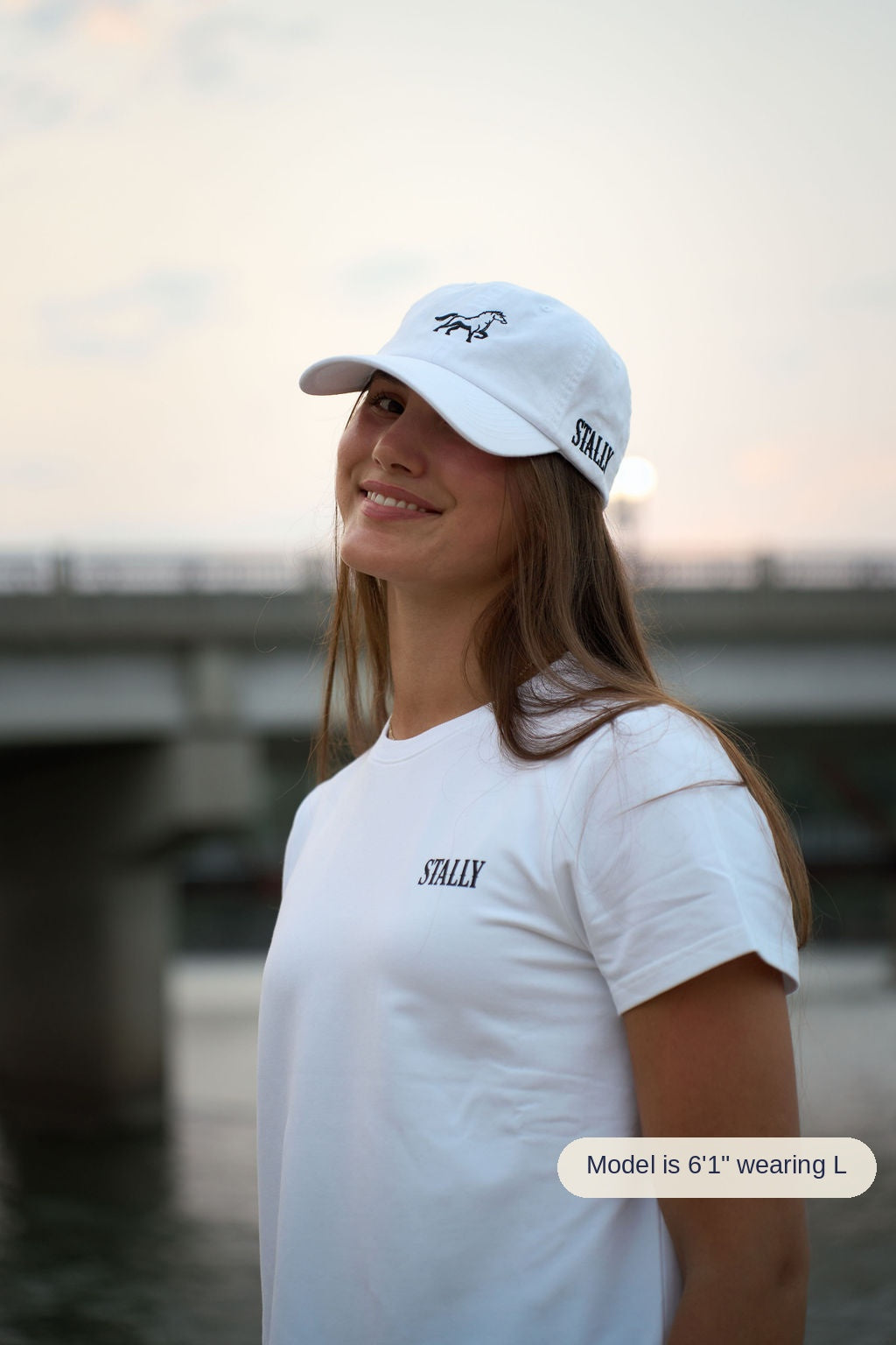 A woman wearing a white crewneck t-shirt and a white baseball cap with black 'STALLY' branding, standing outdoors near a bridge at sunset.