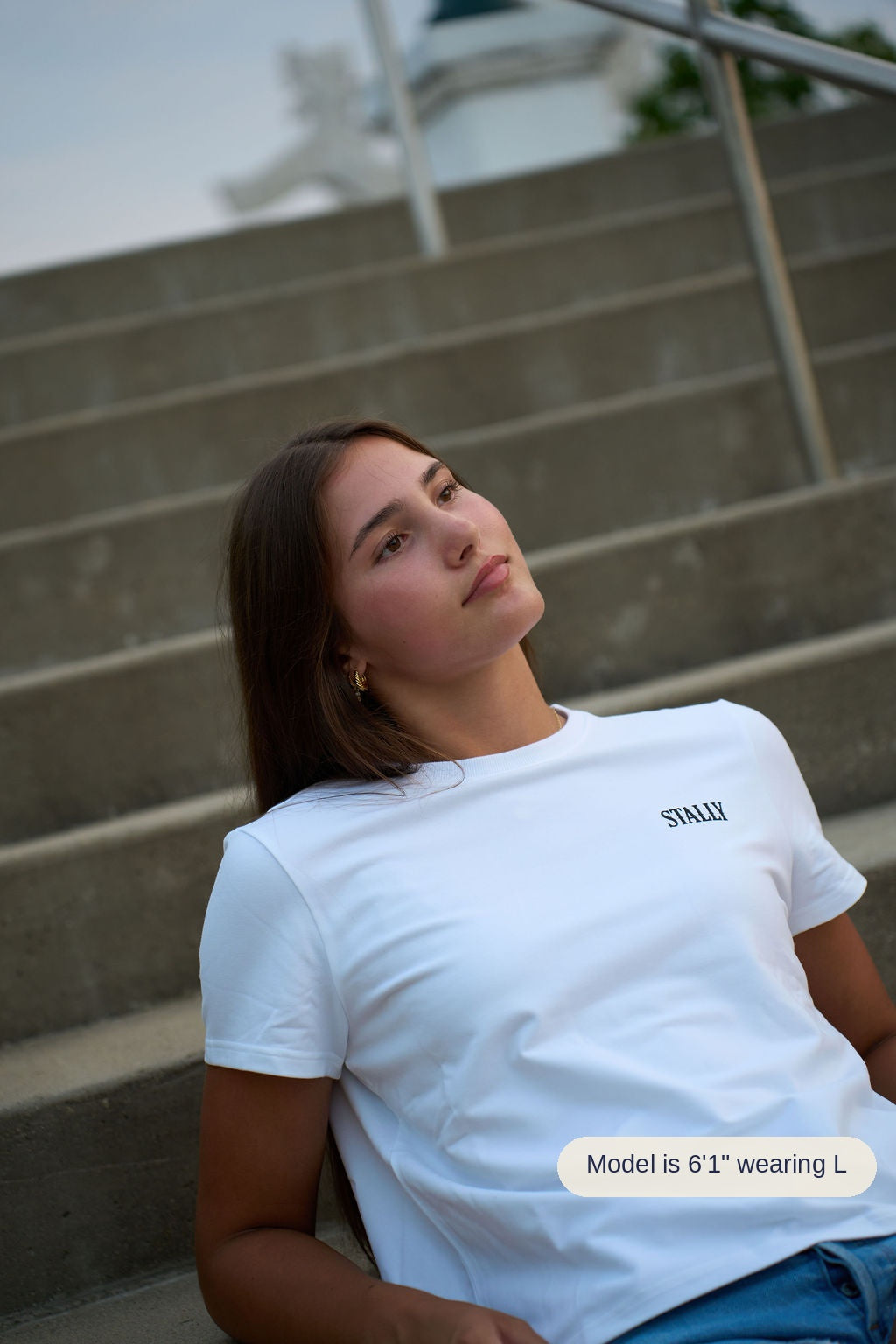 A young woman with brown hair sits on concrete stairs wearing a white crewneck t-shirt with the brand name 'STALLY' printed in black on the left chest, paired with blue jeans.