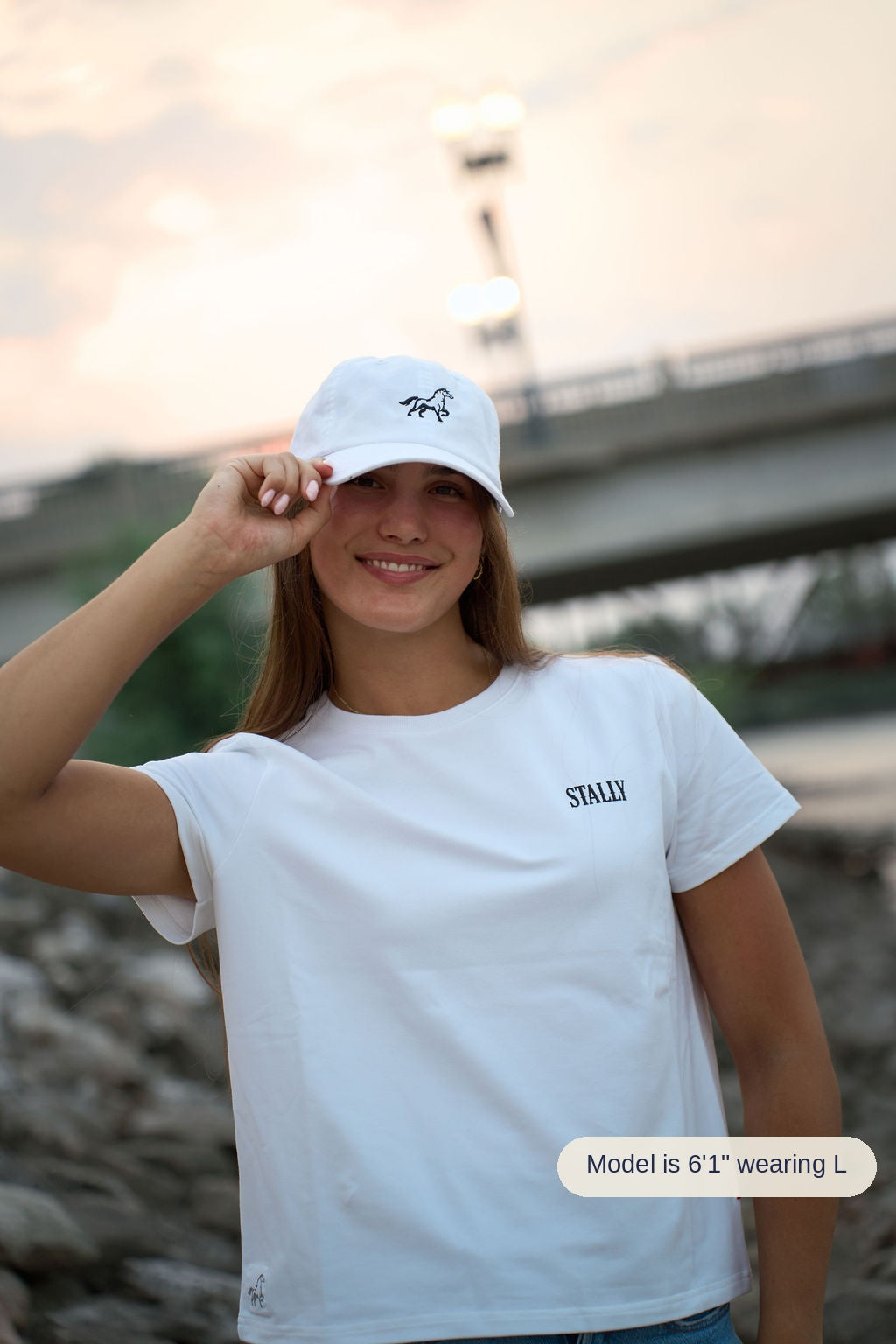 A woman wearing a white crewneck t-shirt with 'STALLY' printed on the chest and a matching white baseball cap, posing outdoors on a rocky shoreline with a bridge in the background.