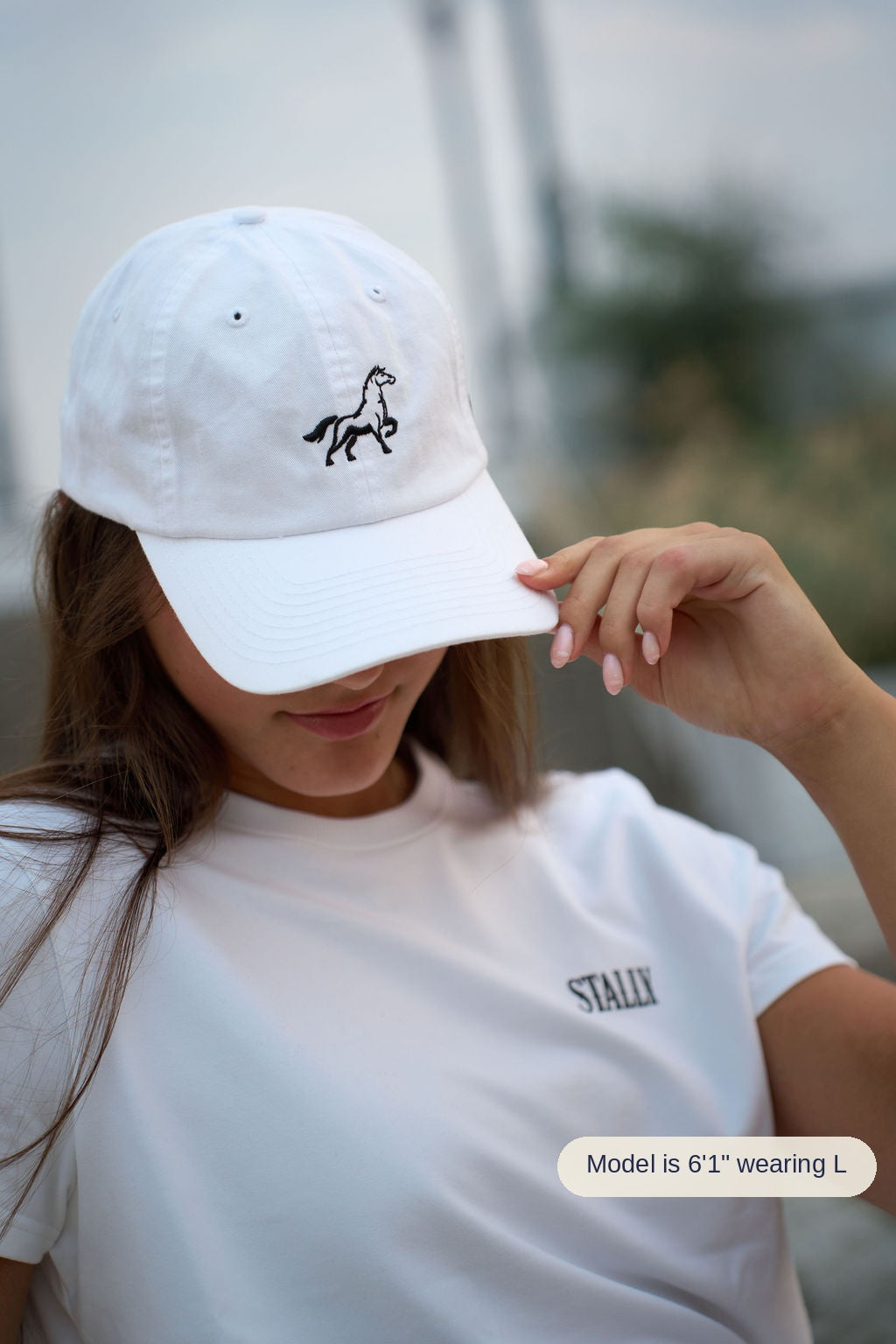 A person wearing a white baseball cap with a black embroidered horse logo and a white T-shirt with a small black 'STALLY' logo on the chest, photographed outdoors with a blurred background.