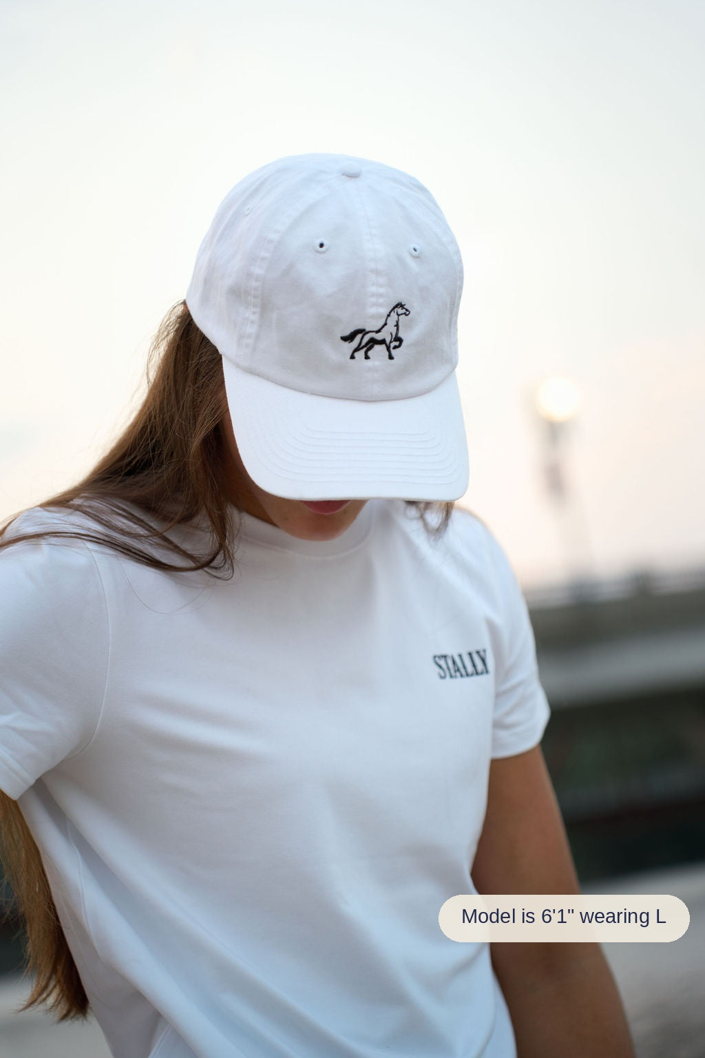 A person wearing a white baseball cap with a black horse logo and a white t-shirt with 'STALLY' printed on the chest, photographed outdoors against a softly blurred background.