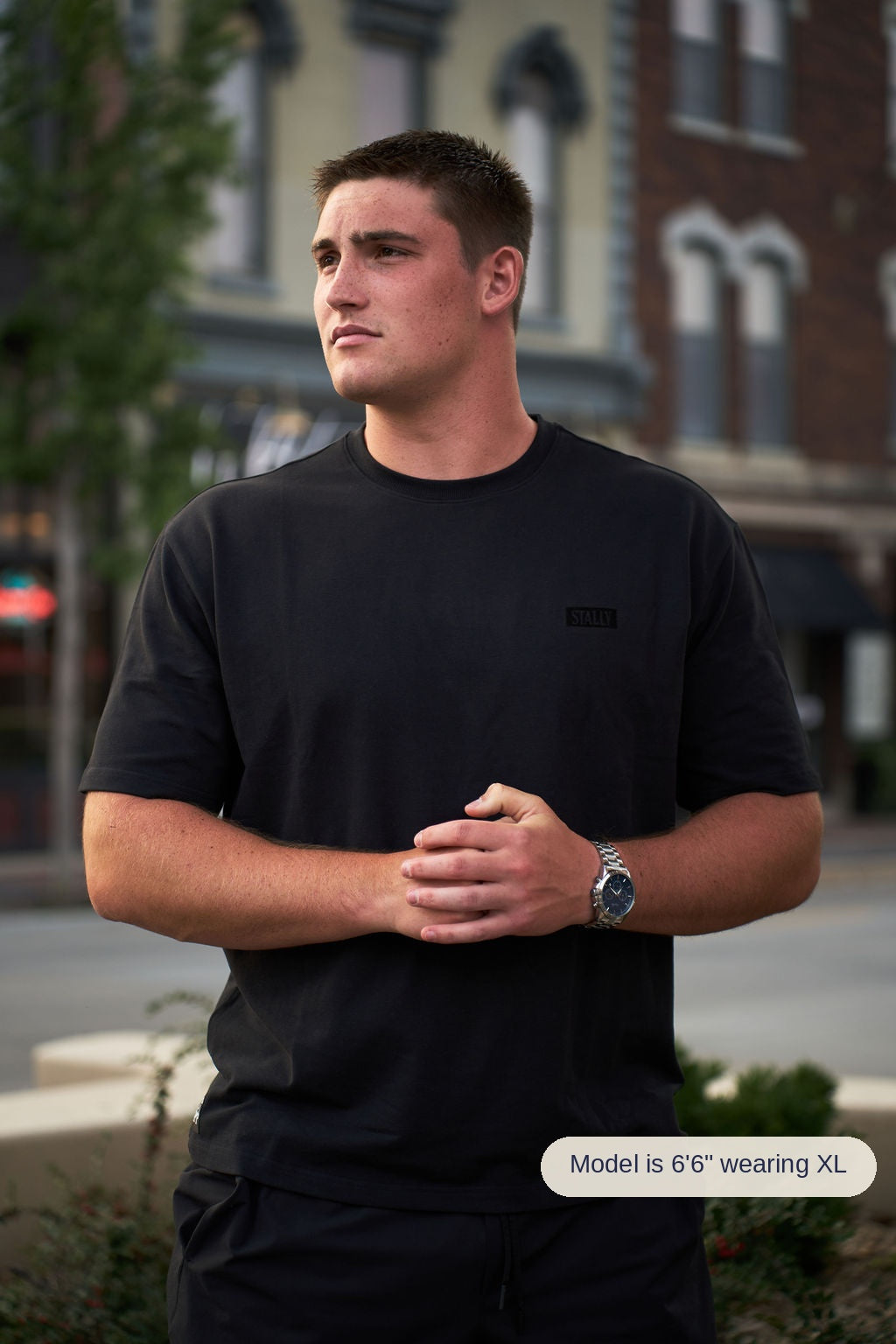 A man wearing a black short-sleeve crewneck t-shirt with a small black rectangular logo patch on the left chest, standing outdoors in front of a blurred brick building.