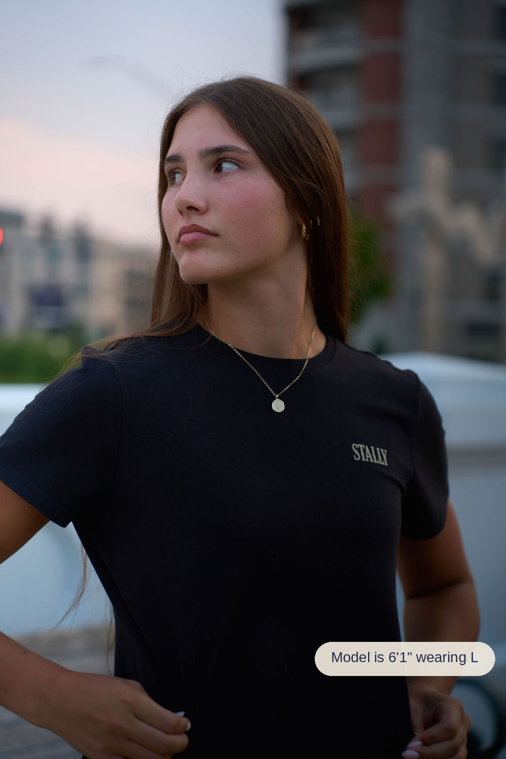 A woman wearing a black crewneck t-shirt with the brand name 'STALLY' printed in white on the left chest, photographed outdoors against a blurred city background at dusk.