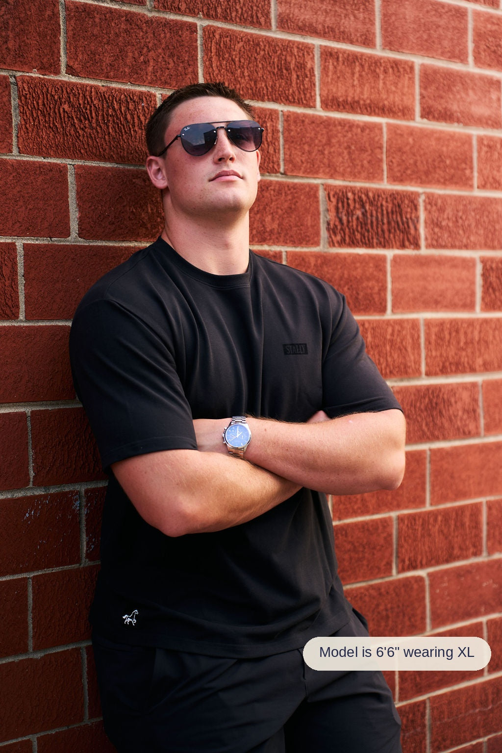 A man wearing a black t-shirt and black pants leans against a red brick wall, wearing dark sunglasses and a silver watch.