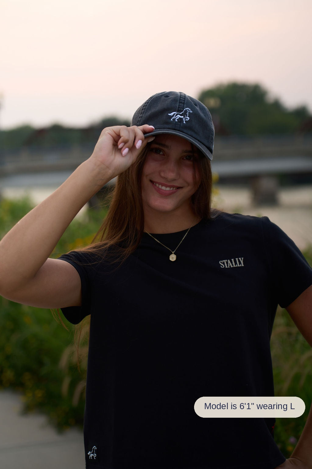 A woman wearing a black crewneck t-shirt with 'STALLY' printed on the chest and a matching black baseball cap, posing outdoors with a blurred background.