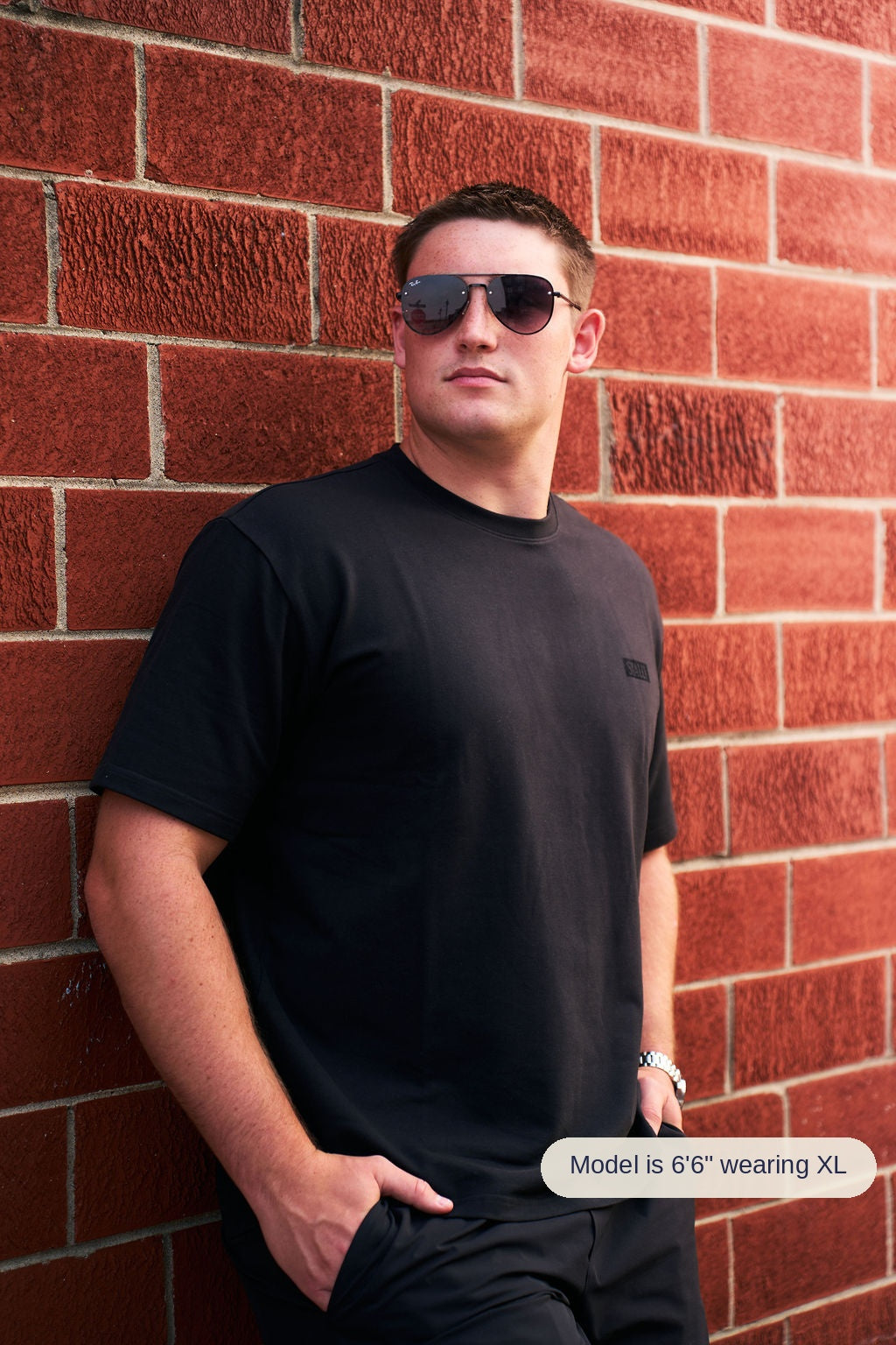 A man wearing a black crewneck t-shirt and black pants, standing against a red brick wall and wearing dark aviator sunglasses.