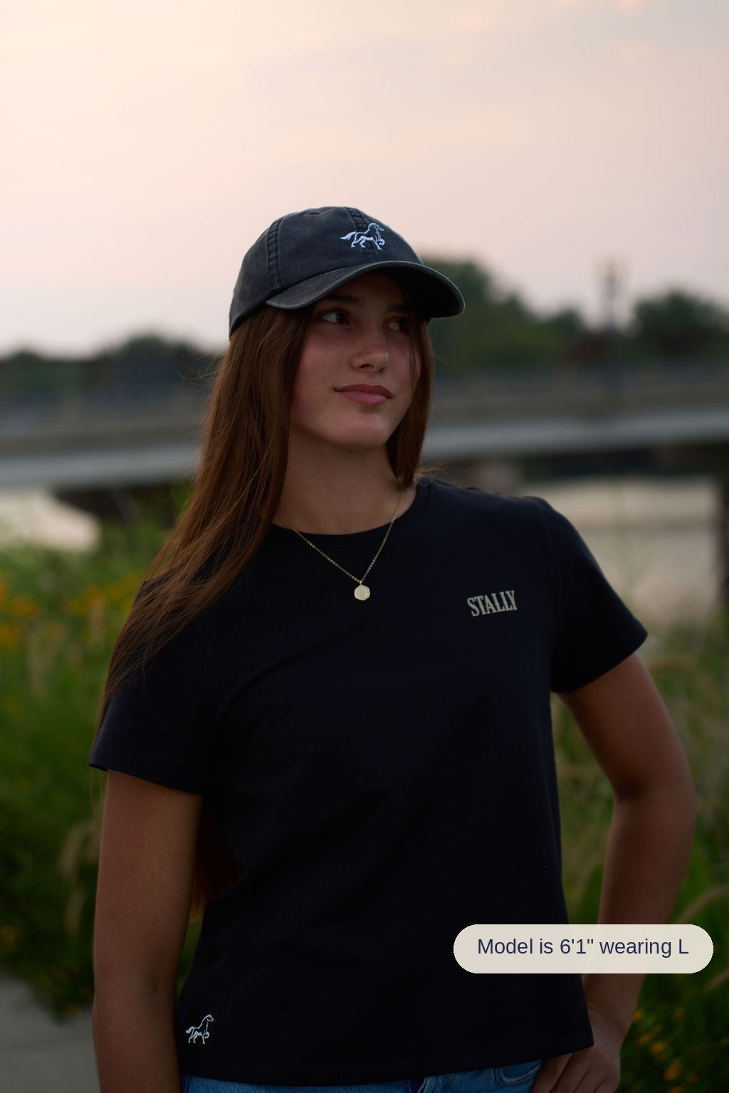 A young woman wearing a black t-shirt with 'STALLY' printed on the chest and a matching black baseball cap, standing outdoors with a blurred background of water and greenery.