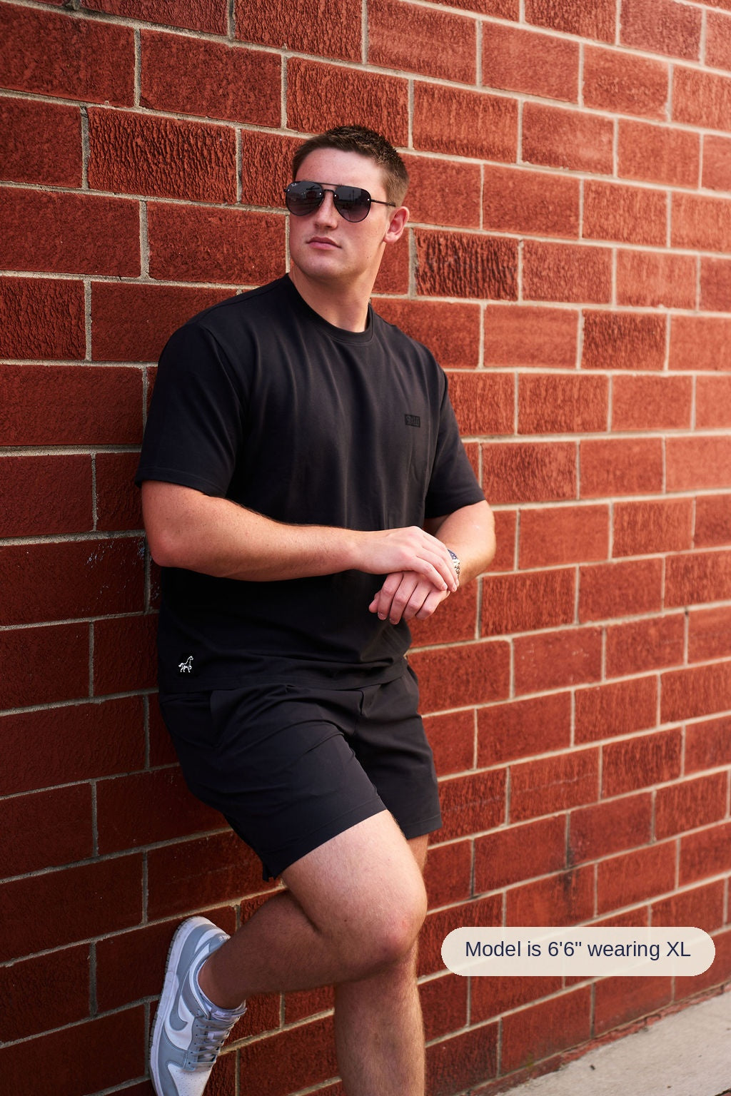 A man wearing a black t-shirt, black shorts, and sunglasses leans against a red brick wall, with one leg bent up. He is wearing white and blue sneakers.