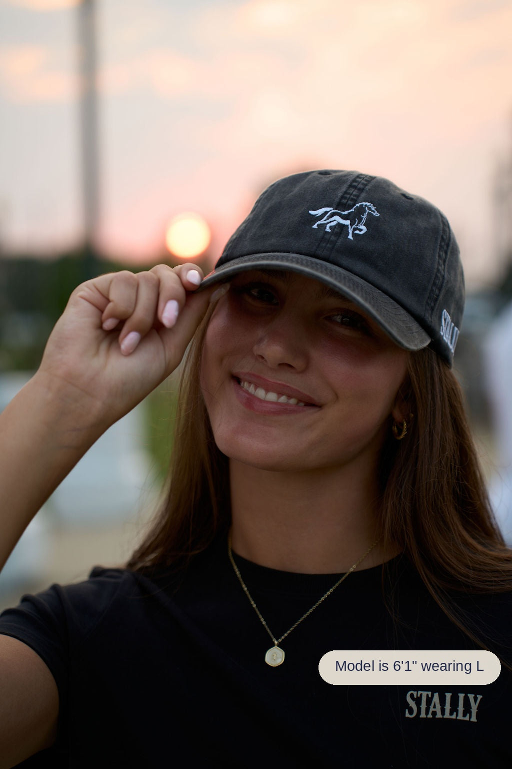 A woman outdoors at sunset wearing a dark washed baseball cap with a white embroidered horse logo and a black t-shirt with 'STALLY' printed on the chest.