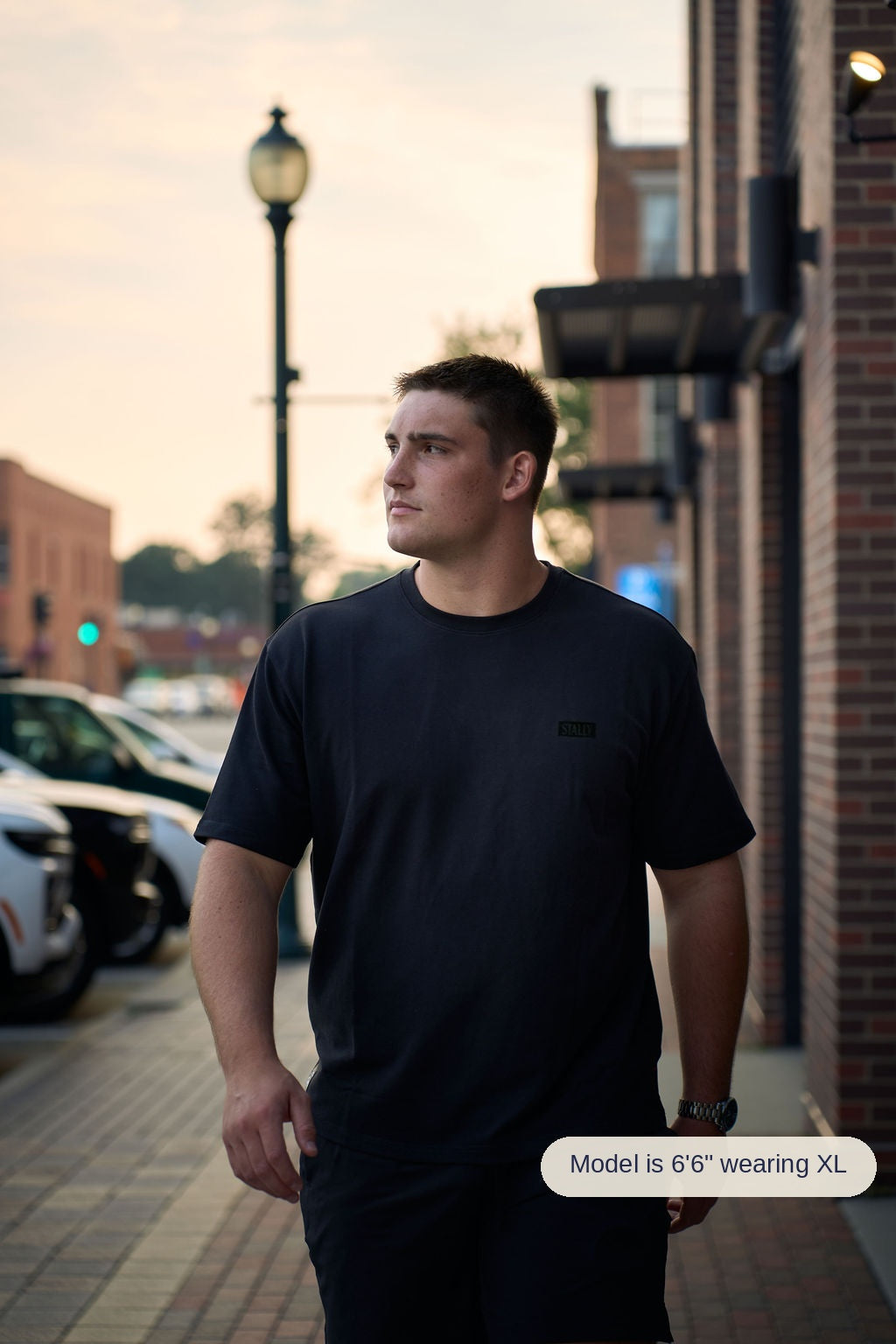A man wearing a black crewneck t-shirt and black shorts walking on a city sidewalk next to a brick building.