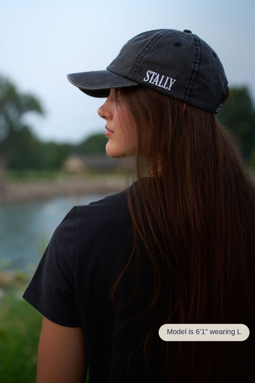 A woman in profile wearing a washed black baseball cap with the word 'STALLY' embroidered in white on the side, photographed outdoors near water with a blurred natural background.