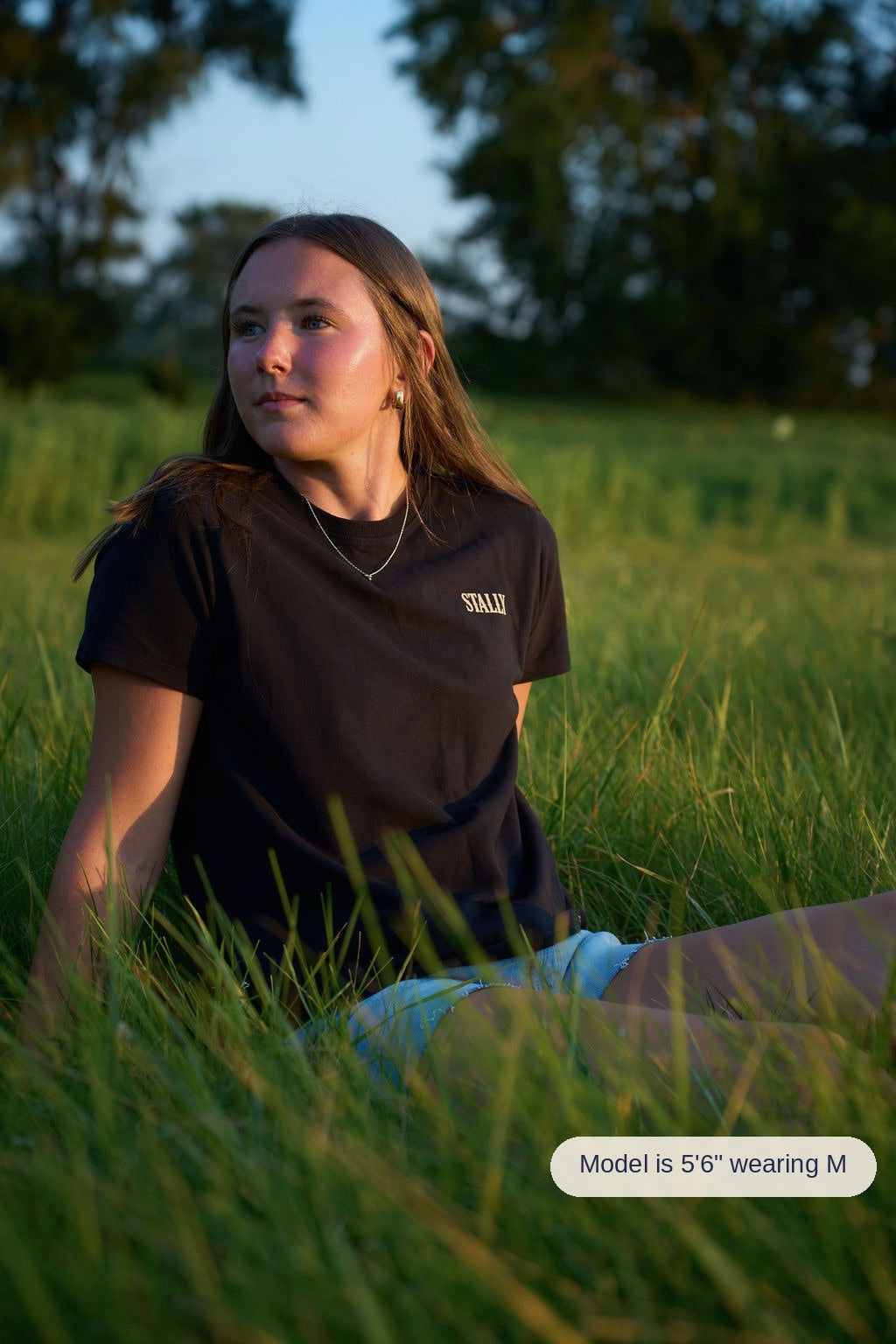 A woman with long brown hair sits in tall green grass at golden hour, wearing a black crewneck t-shirt with 'STALLY' printed in white on the left chest.