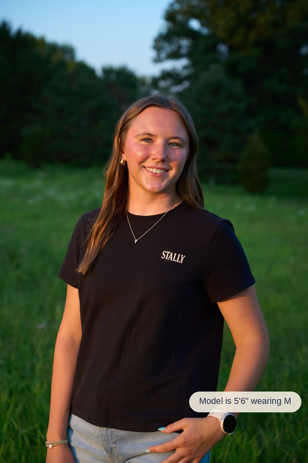A woman with long brown hair stands in a grassy field at sunset wearing a black crewneck t-shirt with 'STALLY' printed in white on the left chest.