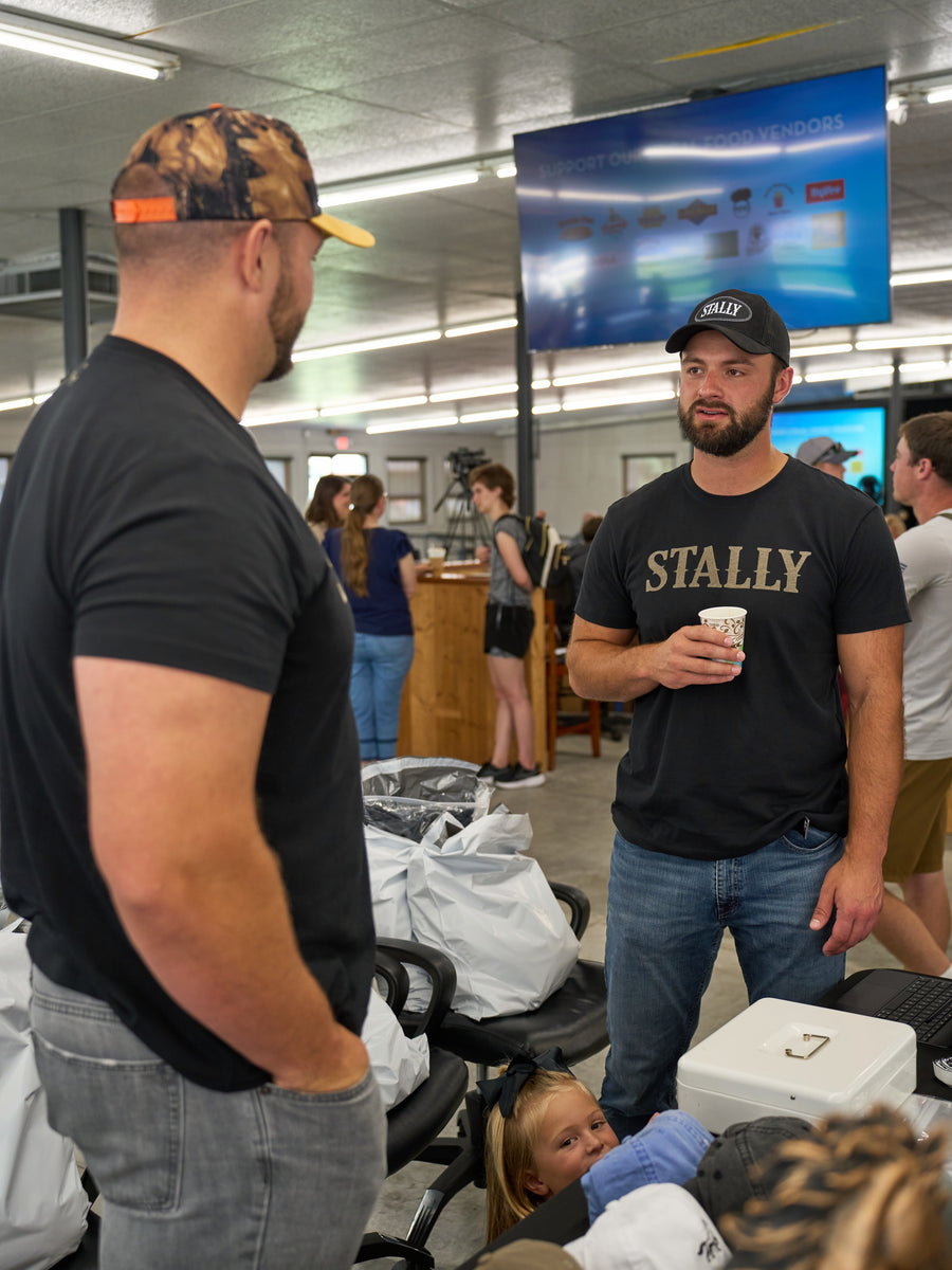 Two men in Stally caps talking beside a merchandise table at a community event.