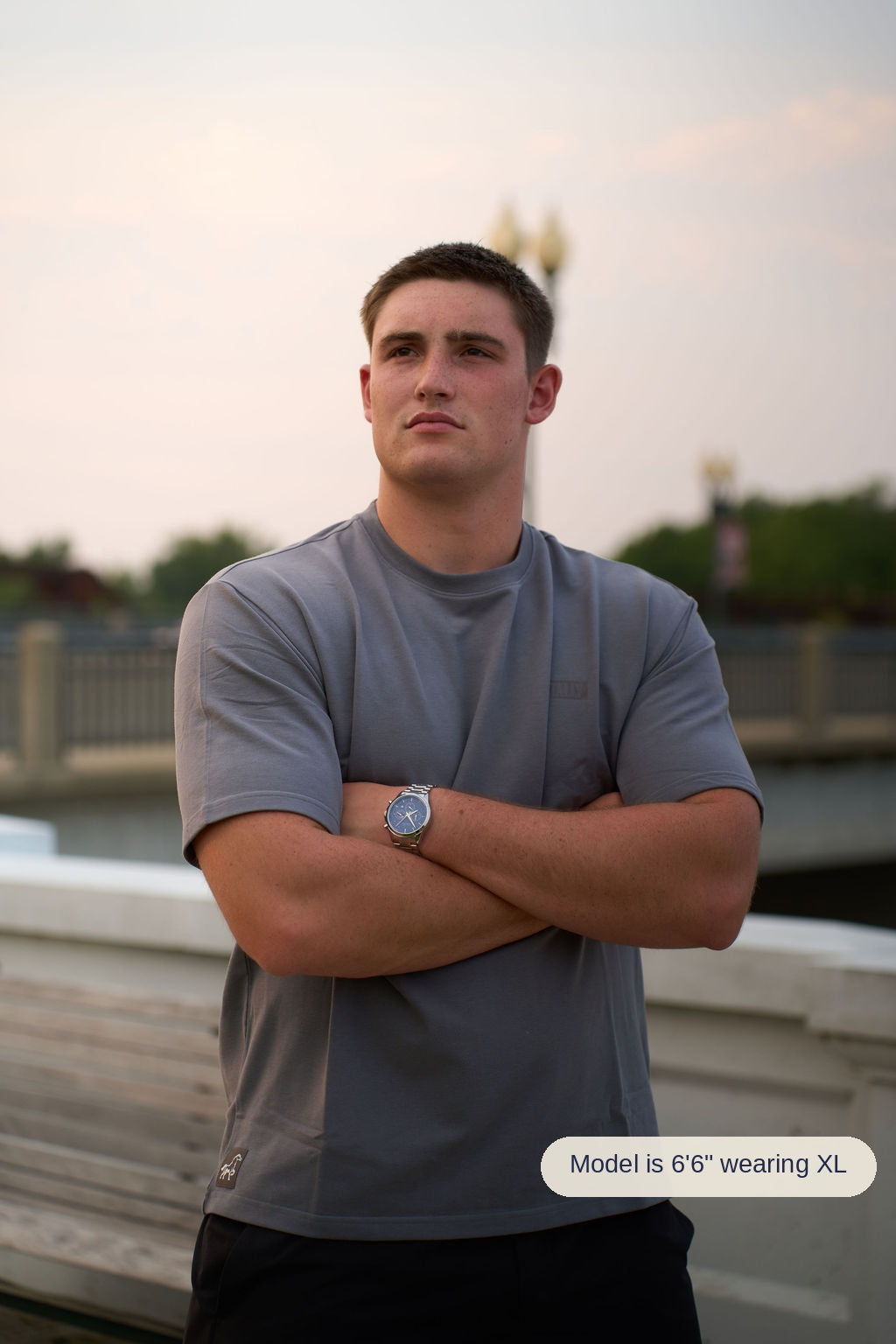 A man wearing a gray athletic t-shirt and black pants stands with his arms crossed, wearing a silver watch on his wrist, set against an outdoor background at dusk.